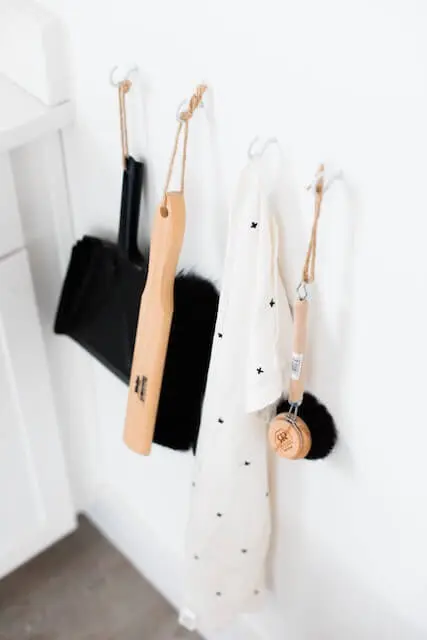 4 cleaning tools, a small brush broom, cloth, a scrubbier and dust pan hanging on a white wall